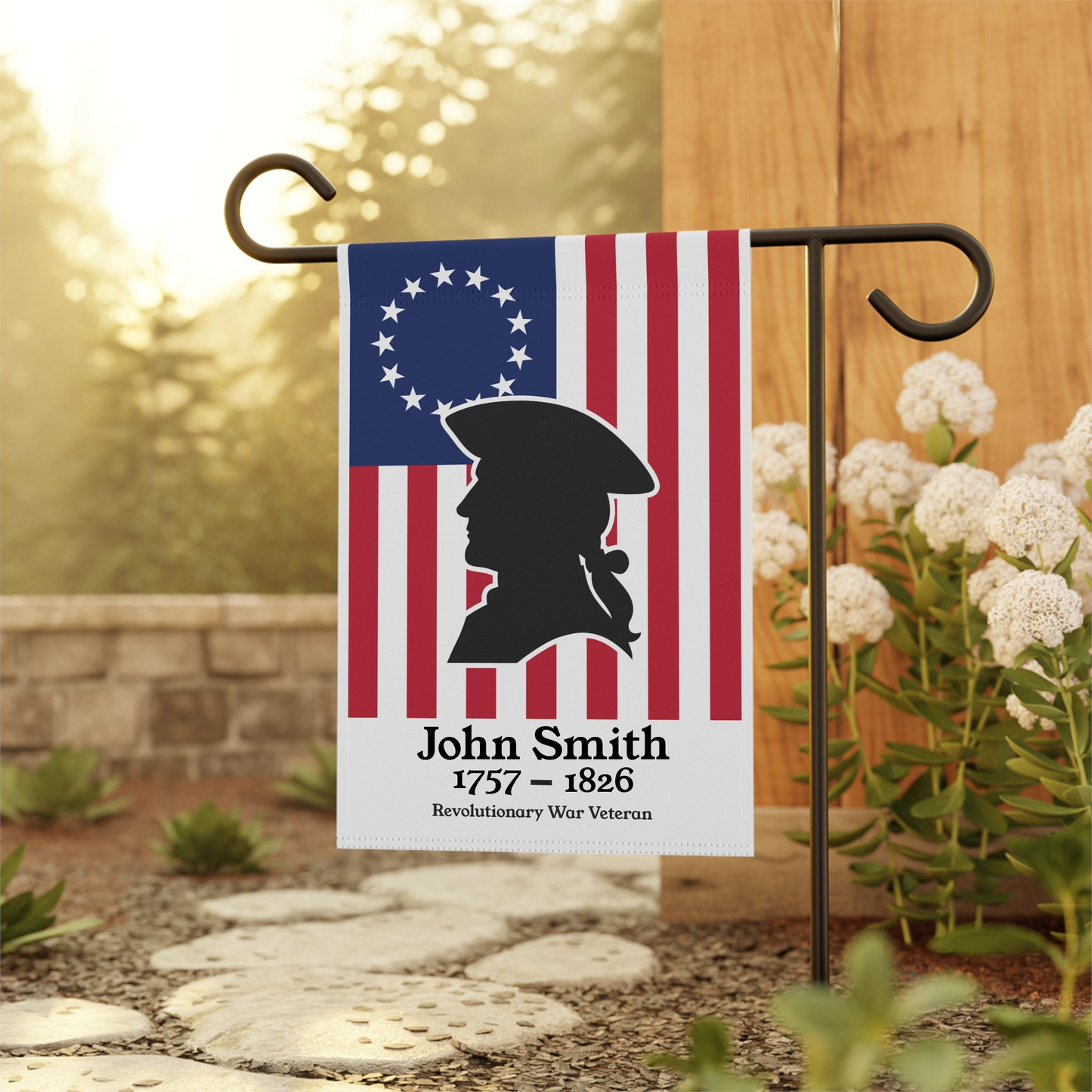 Garden flag with American flag design and silhouette of a Revolutionary War veteran, mounted on a stand outdoors.